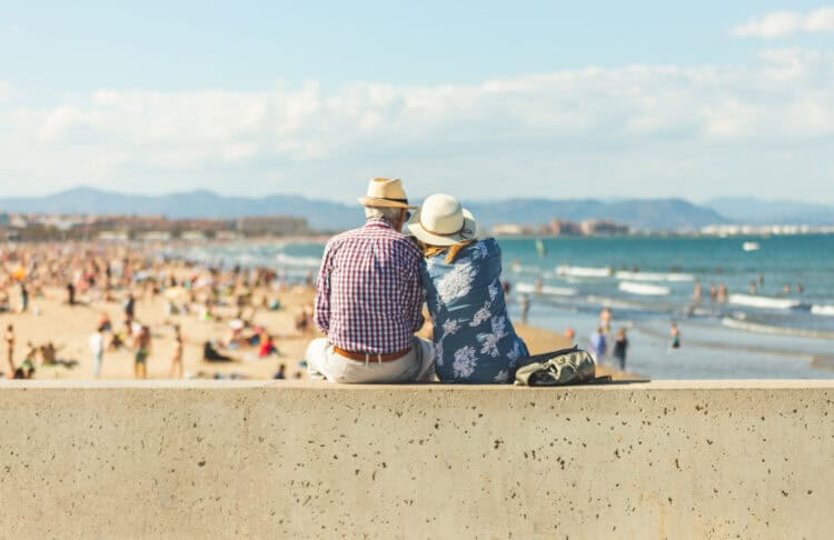 Retired couple wearing summer hats sitting on a concrete wall, overlooking a busy beach with ocean waves, distant mountains, and a partly cloudy sky.