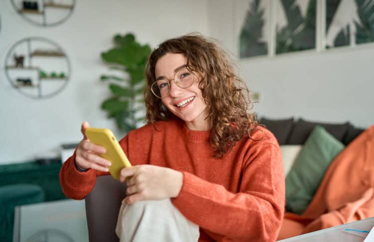 Young adult in an orange sweater holding a yellow smartphone, seated in a cozy living room with a gray couch, green plant, and abstract wall art in the background.